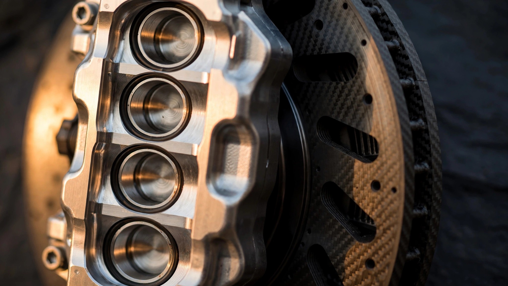 Macro photograph of a multi-piston monobloc brake caliper clamping a carbon ceramic rotor with warm directional lighting