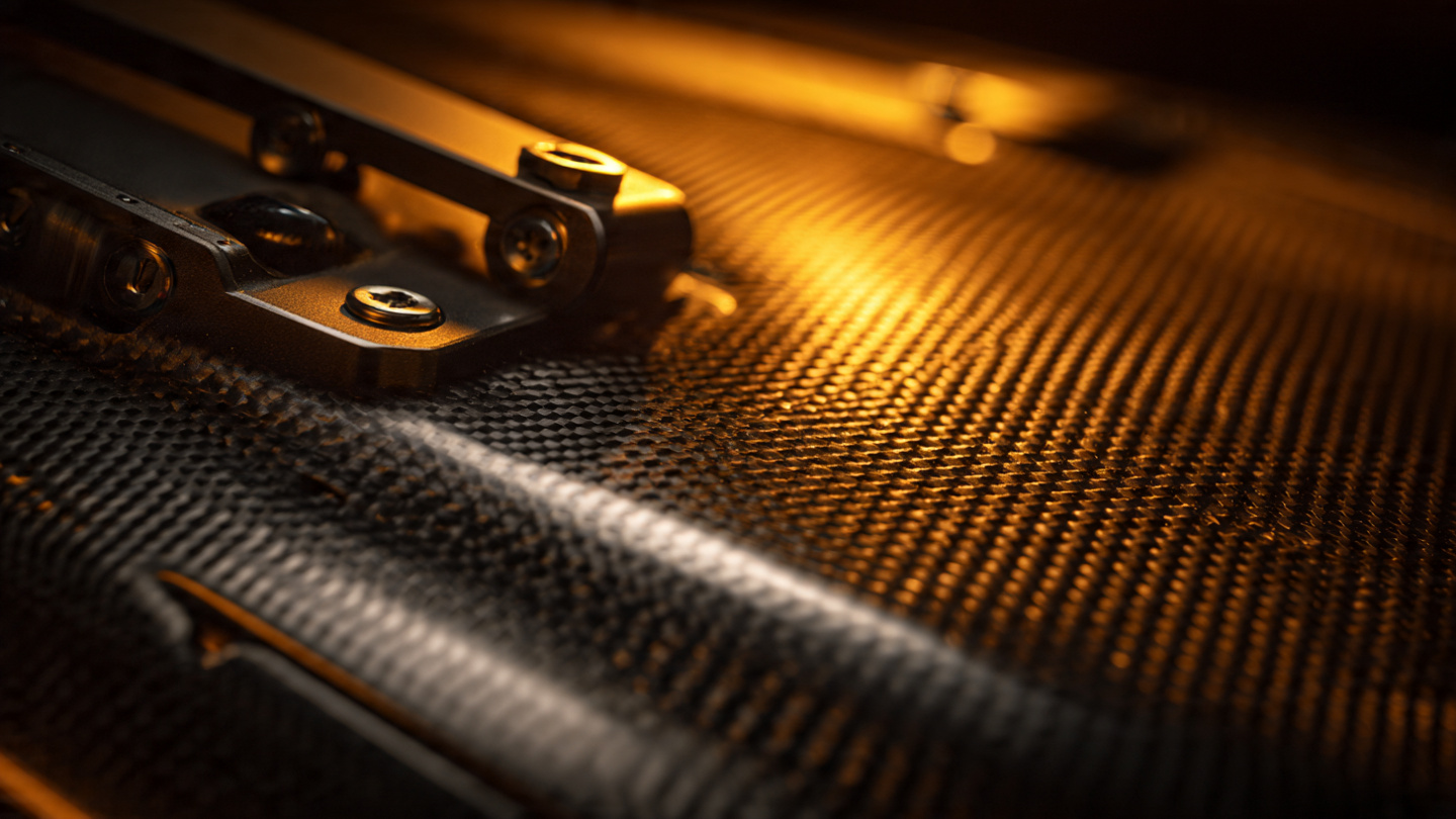Macro photograph of carbon fiber weave with machined metal brackets and hardware under warm amber workshop lighting